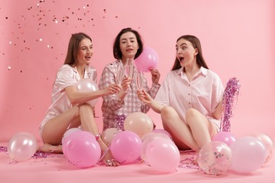 Photo of Pajama party. Happy friends with balloons and tinsel clinking glasses under falling confetti on pink background