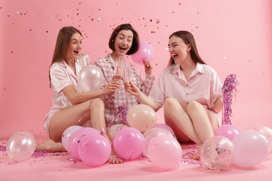 Photo of Pajama party. Happy friends with balloons and tinsel clinking glasses under falling confetti on pink background