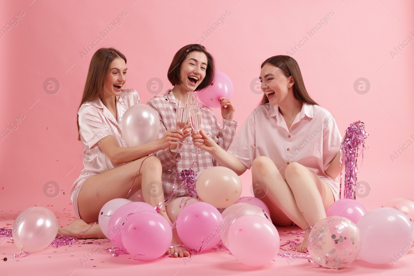 Photo of Pajama party. Happy friends with balloons and tinsel clinking glasses on pink background