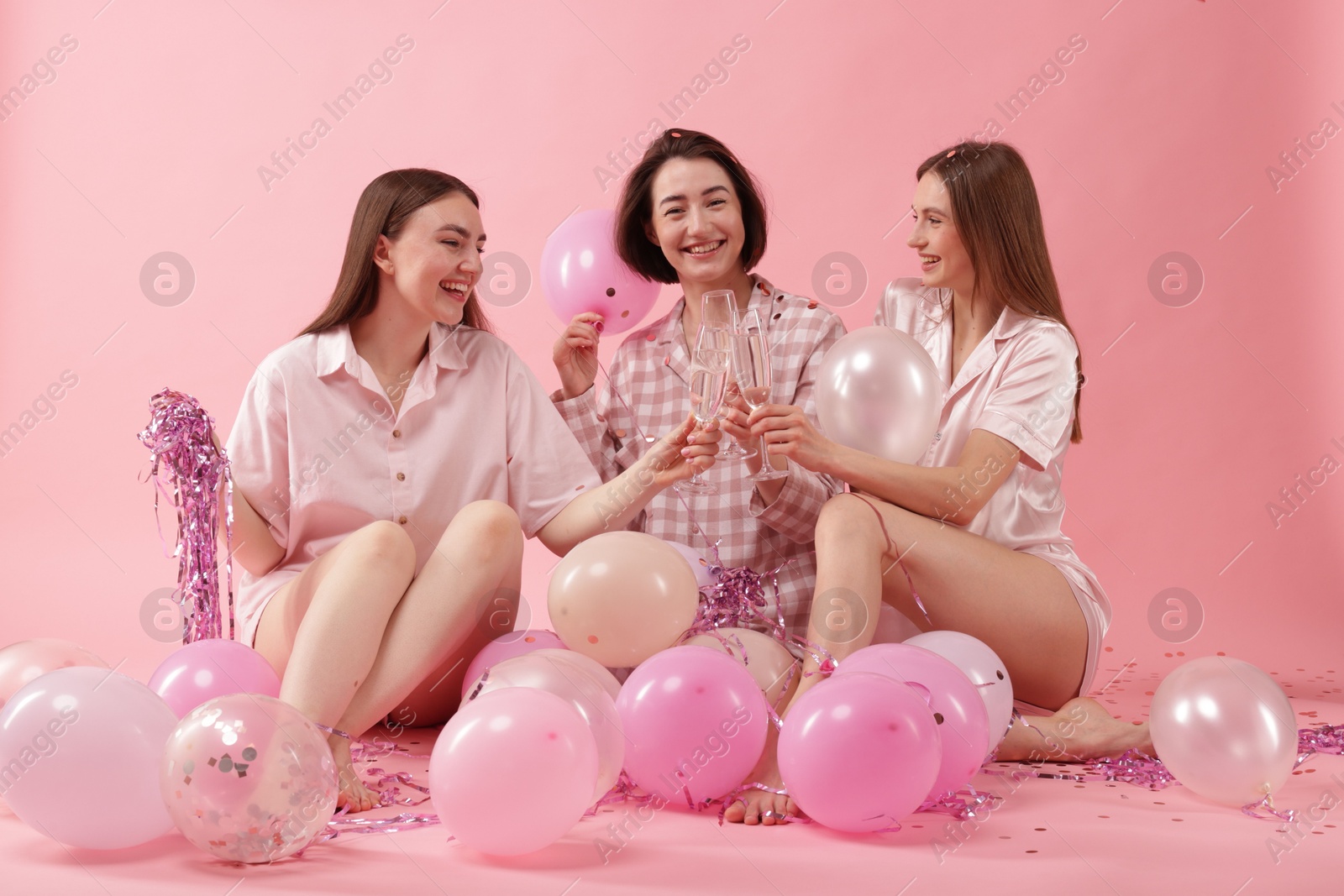 Photo of Pajama party. Happy friends with balloons and tinsel clinking glasses on pink background
