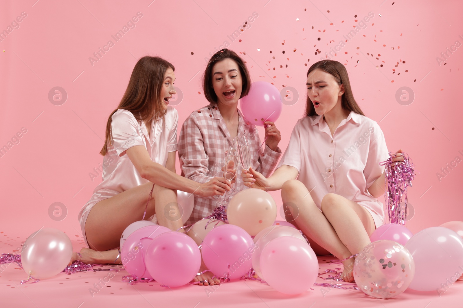 Photo of Pajama party. Happy friends with balloons and tinsel clinking glasses under falling confetti on pink background