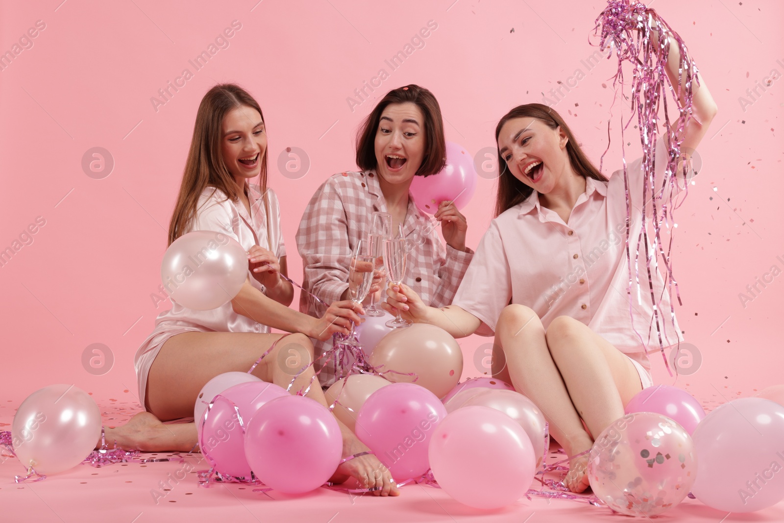 Photo of Pajama party. Happy friends with balloons and tinsel clinking glasses under falling confetti on pink background