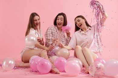 Photo of Pajama party. Happy friends with balloons and tinsel clinking glasses under falling confetti on pink background