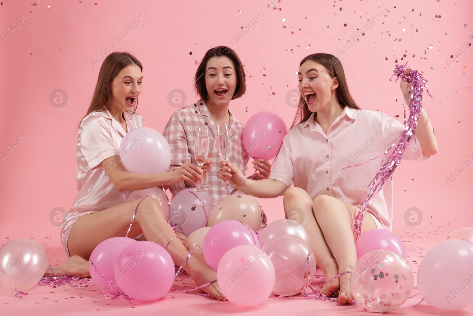 Photo of Pajama party. Happy friends with balloons and tinsel clinking glasses under falling confetti on pink background