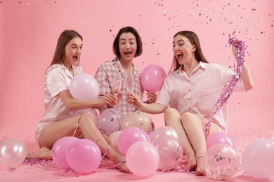 Photo of Pajama party. Happy friends with balloons and tinsel clinking glasses under falling confetti on pink background