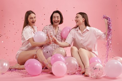 Photo of Pajama party. Happy friends with balloons and tinsel clinking glasses under falling confetti on pink background