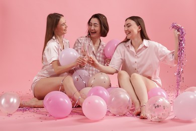 Photo of Pajama party. Happy friends with balloons and tinsel clinking glasses on pink background