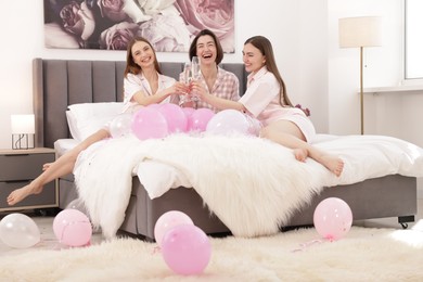 Photo of Pajama party. Happy friends with balloons clinking glasses on bed at home