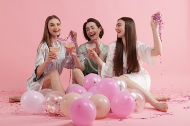 Photo of Pajama party. Happy friends with tinsel and balloons clinking glasses on pink background