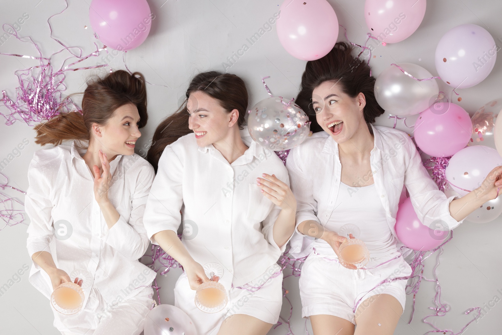 Photo of Pajama party. Happy friends with glasses of alcoholic drink, balloons and tinsel on grey background, top view