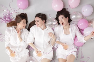 Photo of Pajama party. Happy friends with glasses of alcoholic drink, balloons and tinsel on grey background, top view