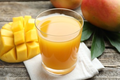 Photo of Tasty mango juice in glass, fresh fruits and green leaves on wooden table, closeup