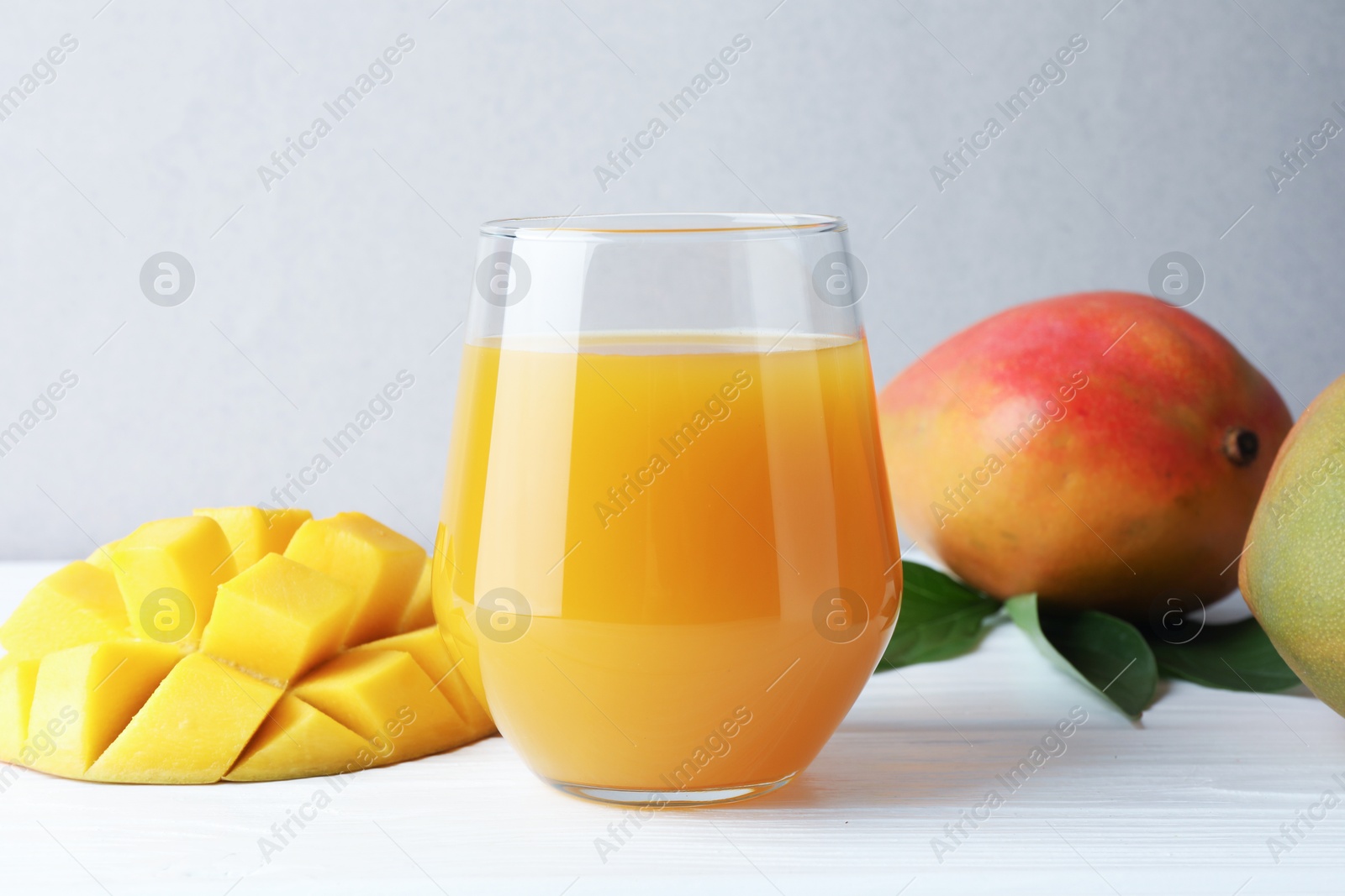 Photo of Tasty mango juice in glass, fresh fruits and green leaves on white wooden table, closeup