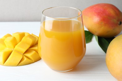 Photo of Tasty mango juice in glass, fresh fruits and green leaves on white wooden table, closeup