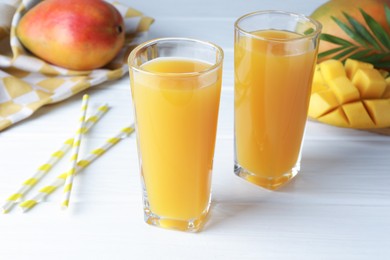 Photo of Tasty mango juice in glasses, fresh fruits, straws and green leaves on white wooden table, closeup