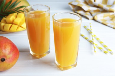 Photo of Tasty mango juice in glasses, fresh fruits, straws and green leaves on white wooden table, closeup