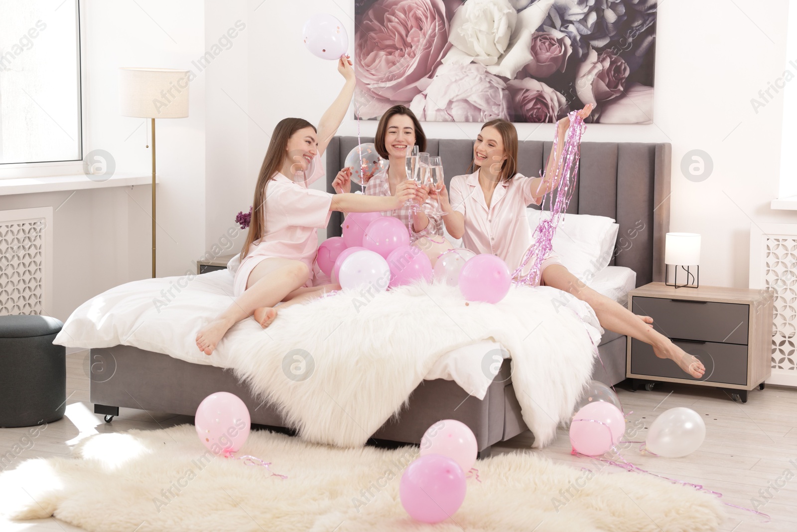 Photo of Pajama party. Happy friends with balloons and tinsel clinking glasses on bed at home