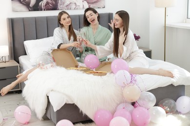 Photo of Pajama party. Happy friends with pizza clinking glasses on bed at home