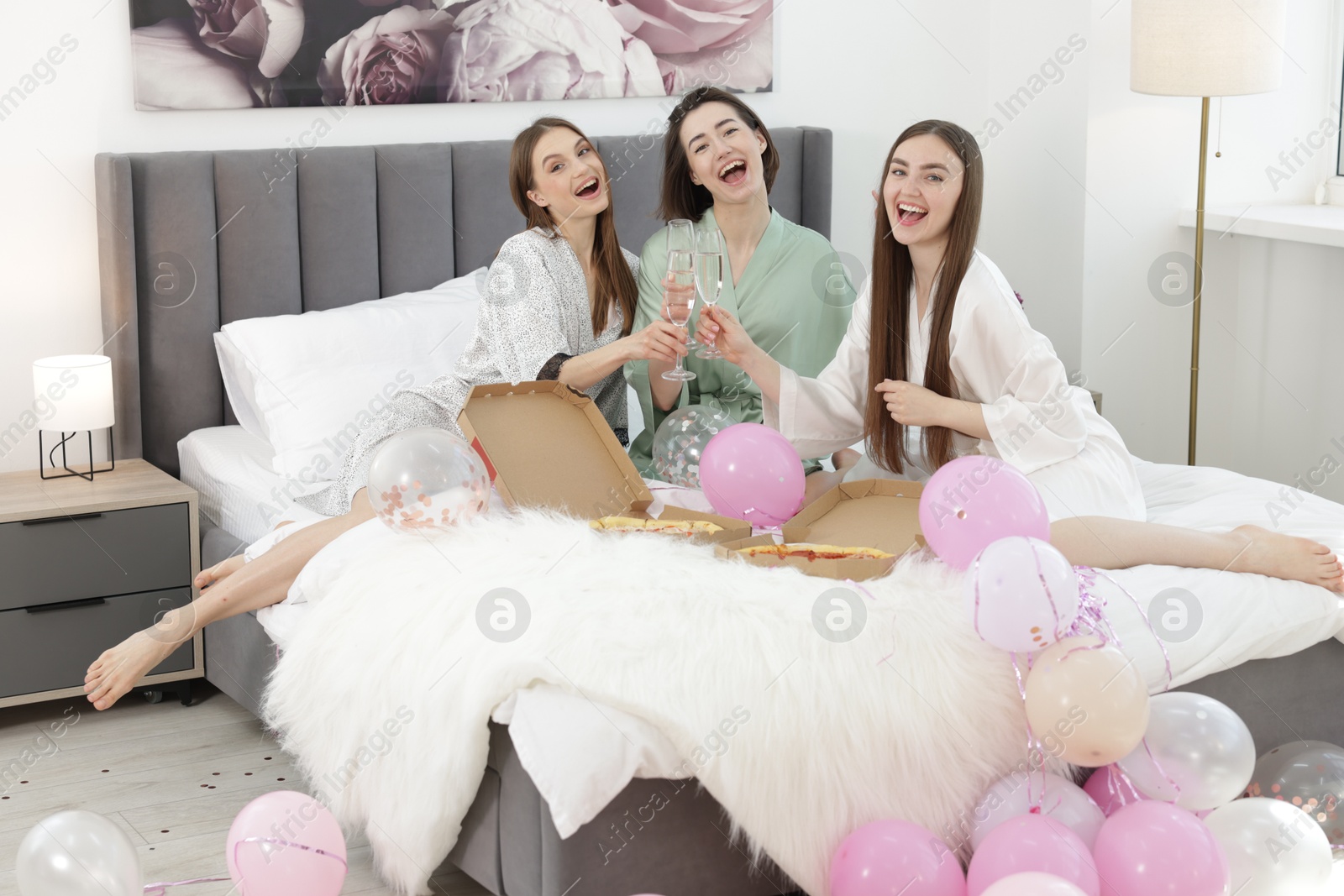 Photo of Pajama party. Happy friends with pizza clinking glasses on bed at home