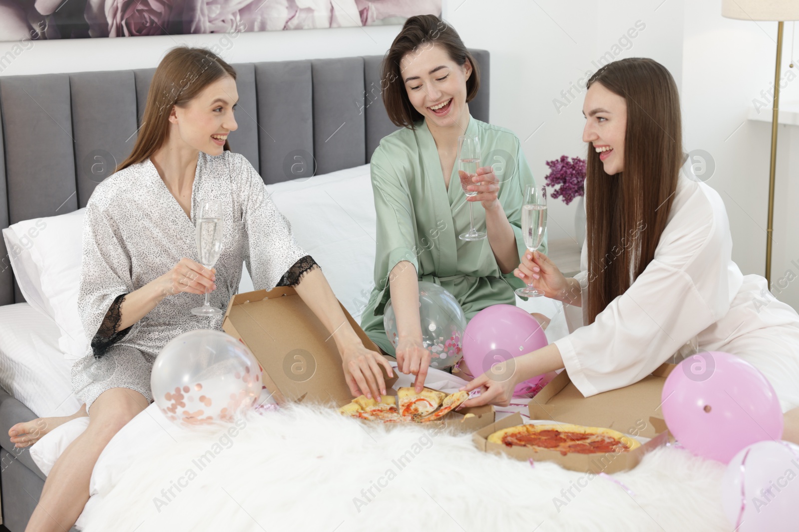 Photo of Pajama party. Happy friends with glasses eating pizza on bed at home