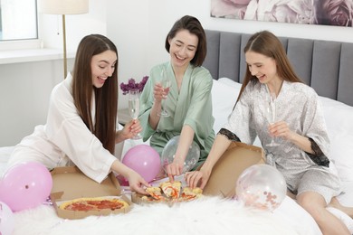 Photo of Pajama party. Happy friends with glasses eating pizza on bed at home