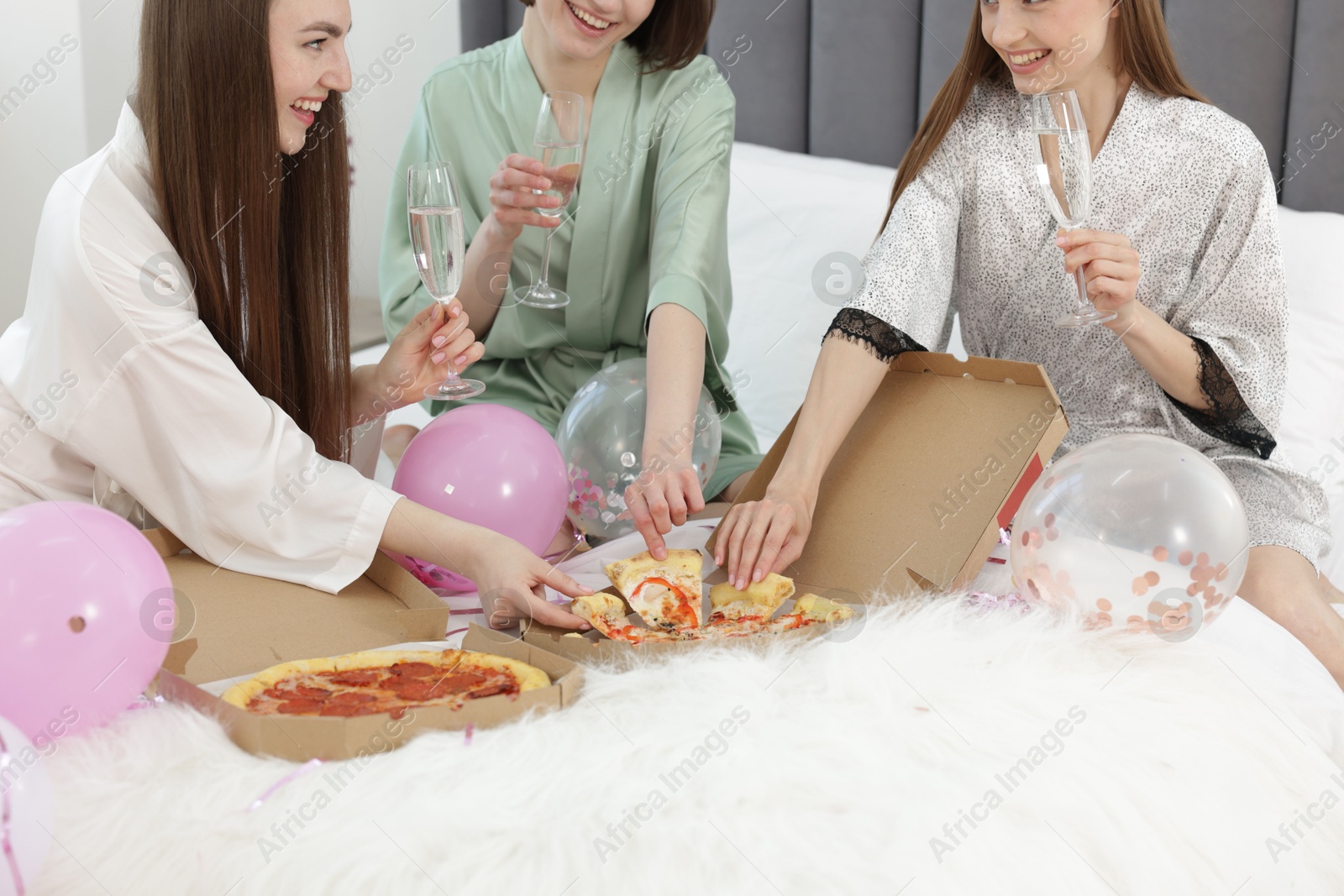 Photo of Pajama party. Happy friends with glasses eating pizza on bed at home, closeup