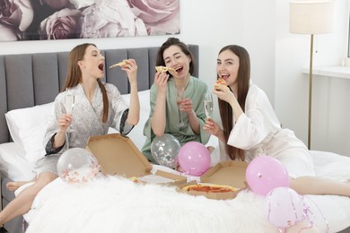 Photo of Pajama party. Happy friends with glasses eating pizza on bed at home