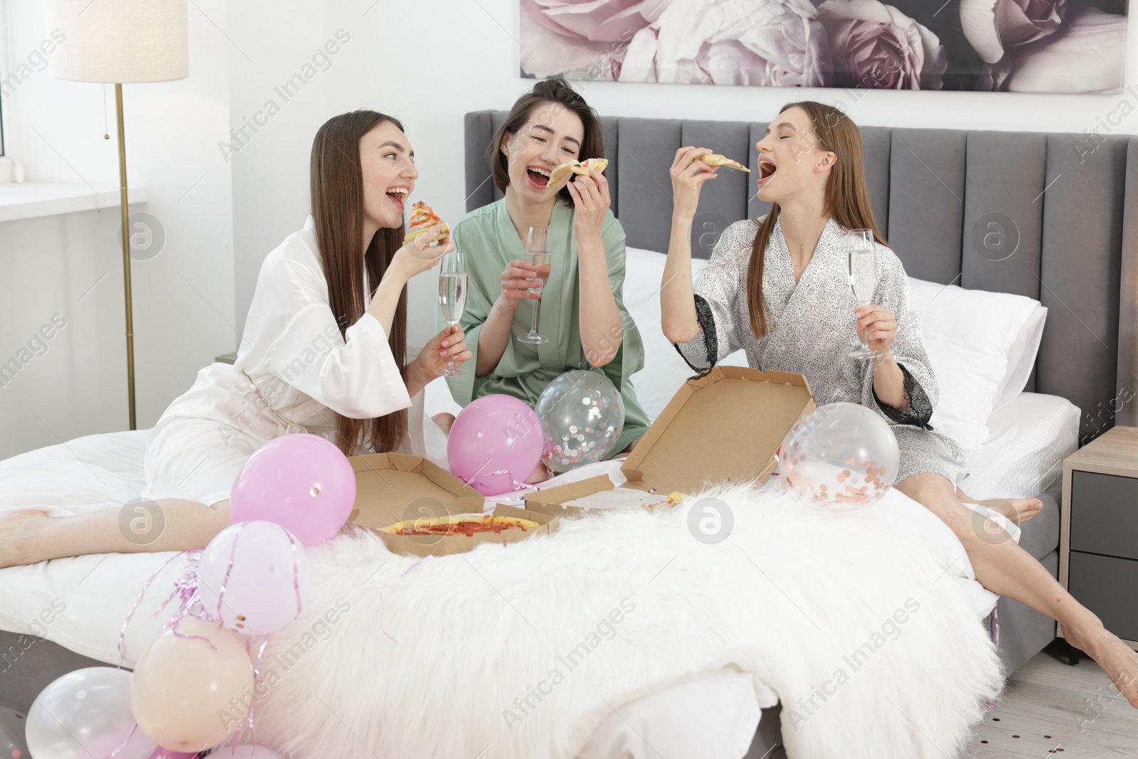 Photo of Pajama party. Happy friends with glasses eating pizza on bed at home