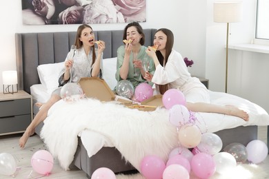 Photo of Pajama party. Happy friends with glasses eating pizza on bed at home