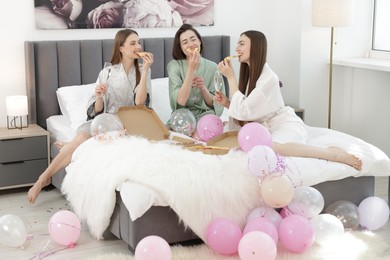 Photo of Pajama party. Happy friends with glasses eating pizza on bed at home