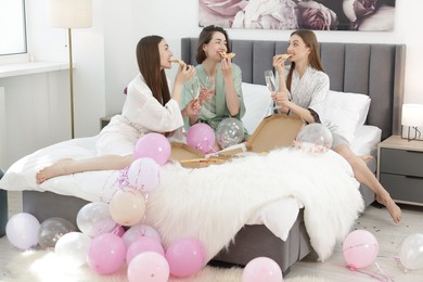 Photo of Pajama party. Happy friends with glasses eating pizza on bed at home