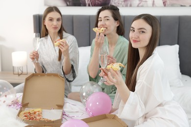 Photo of Pajama party. Happy friends with glasses eating pizza on bed at home