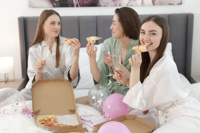 Photo of Pajama party. Happy friends with glasses eating pizza on bed at home