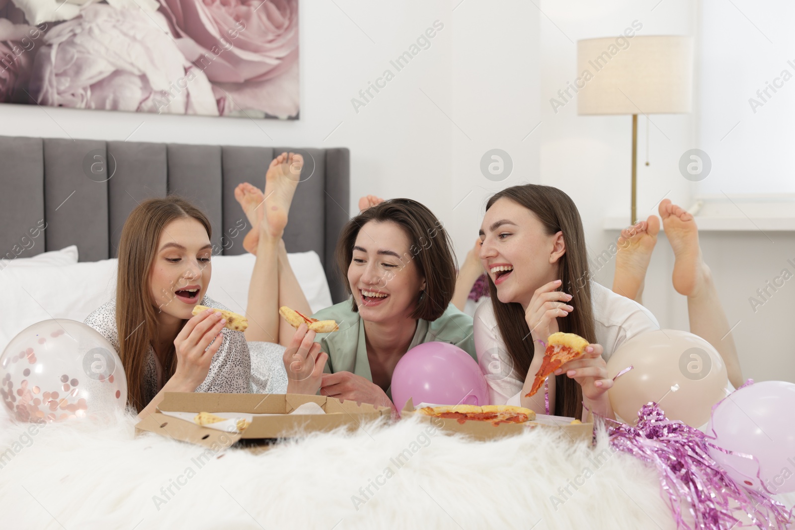 Photo of Pajama party. Happy friends eating pizza on bed at home