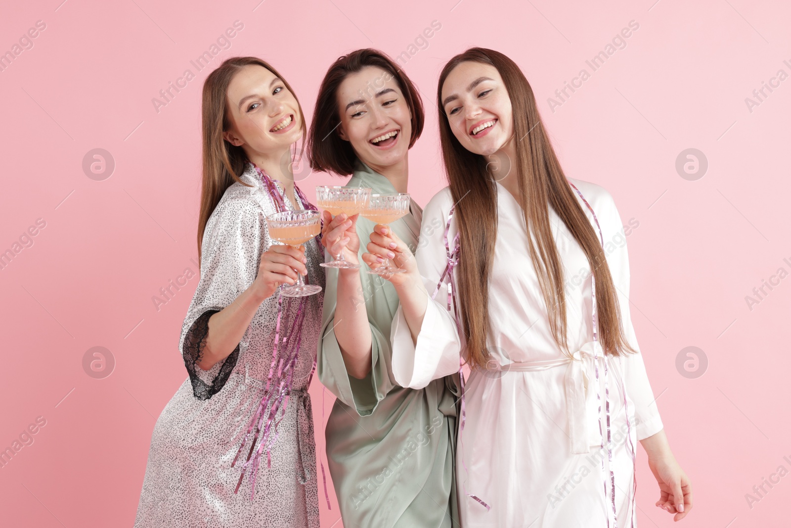 Photo of Pajama party. Happy friends with tinsel clinking glasses on pink background