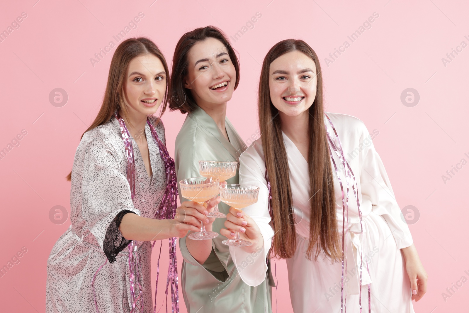 Photo of Pajama party. Happy friends with tinsel clinking glasses on pink background