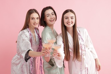 Photo of Pajama party. Happy friends with tinsel clinking glasses on pink background
