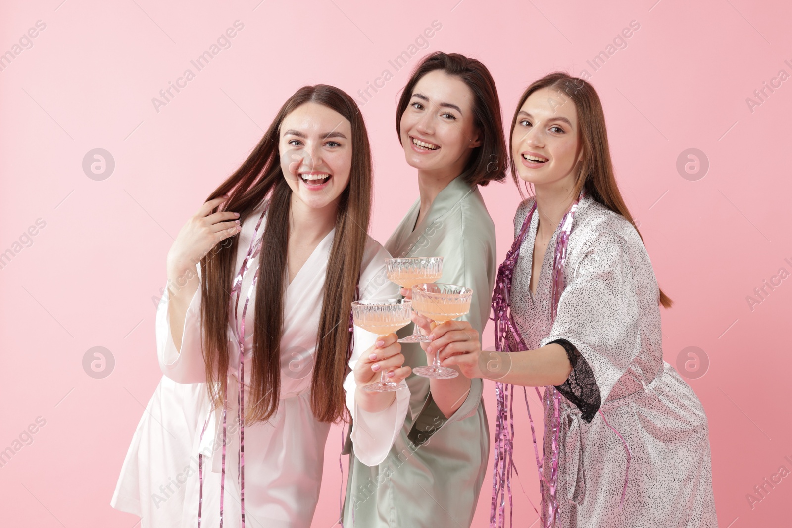 Photo of Pajama party. Happy friends with tinsel clinking glasses on pink background