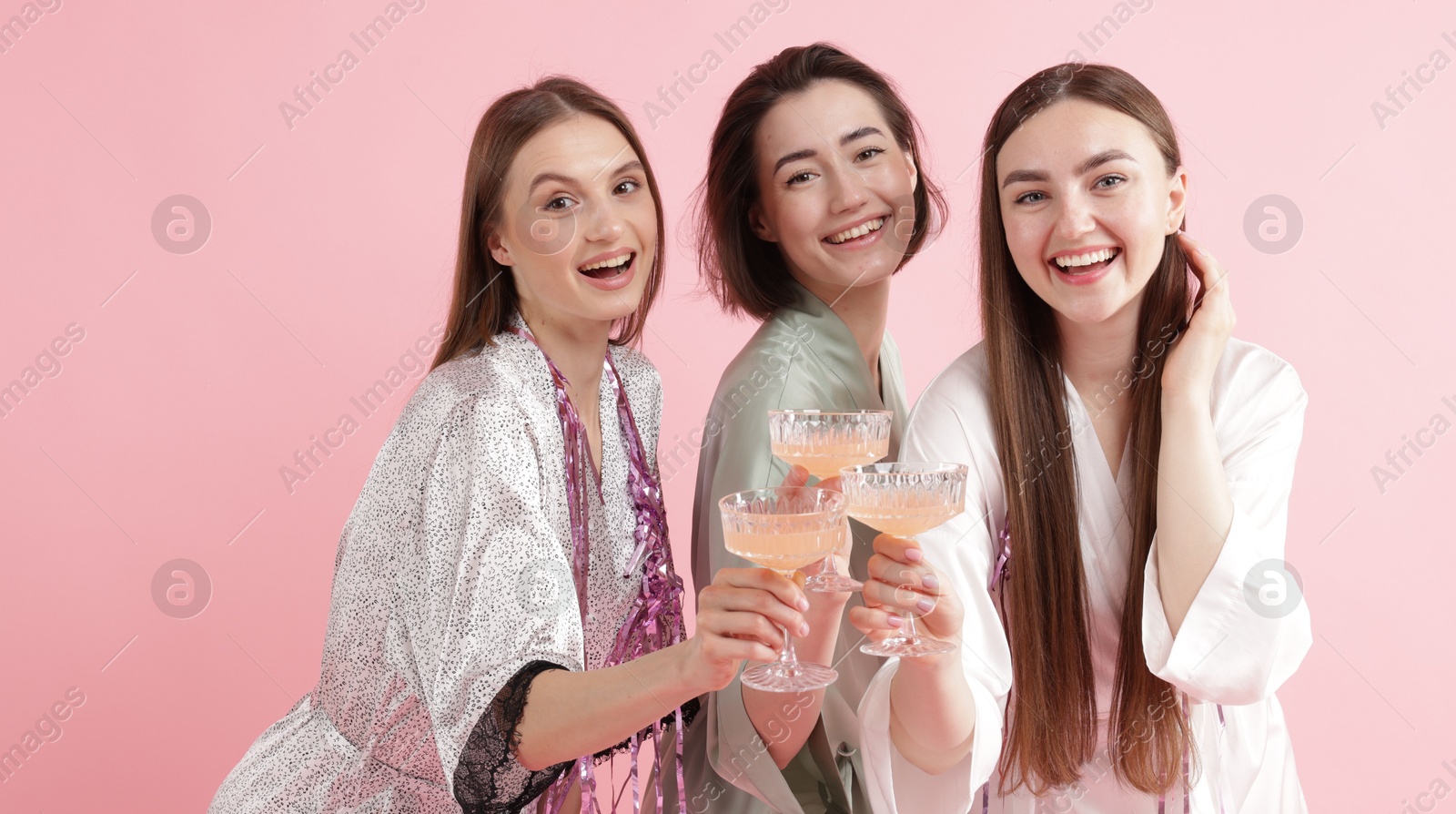 Photo of Pajama party. Happy friends with tinsel clinking glasses on pink background