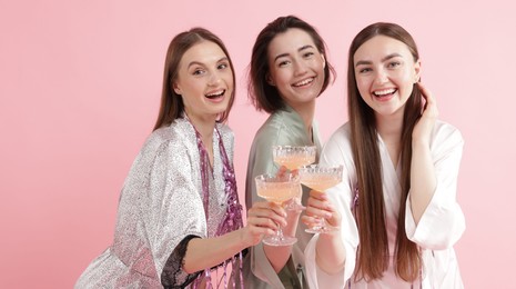Photo of Pajama party. Happy friends with tinsel clinking glasses on pink background