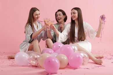 Photo of Pajama party. Happy friends with tinsel and balloons clinking glasses on pink background