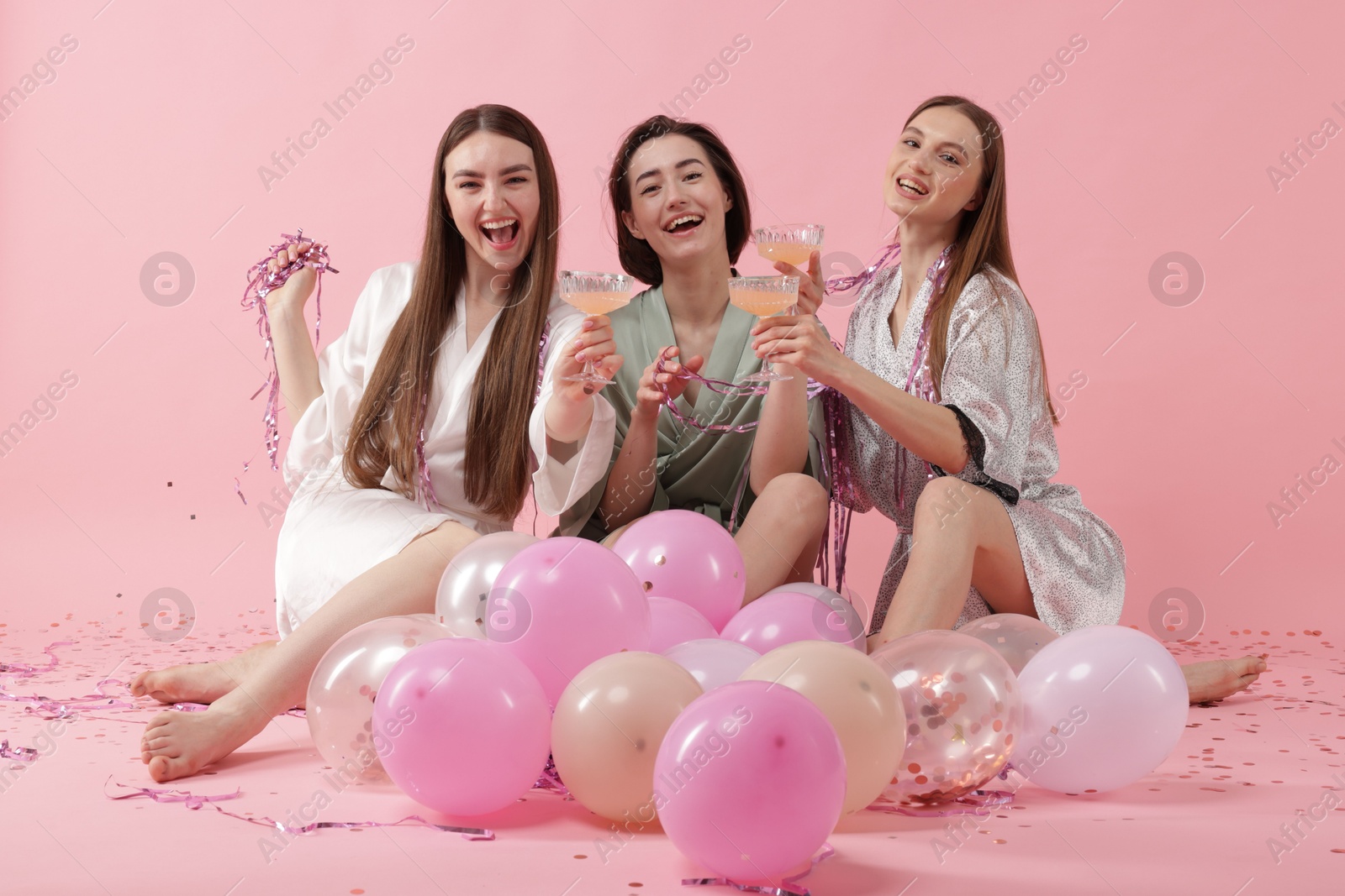 Photo of Pajama party. Happy friends with tinsel and balloons clinking glasses on pink background