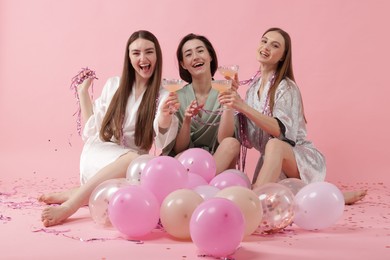 Photo of Pajama party. Happy friends with tinsel and balloons clinking glasses on pink background