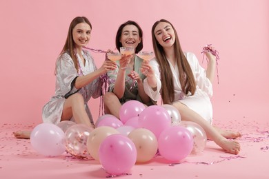 Photo of Pajama party. Happy friends with tinsel and balloons clinking glasses on pink background