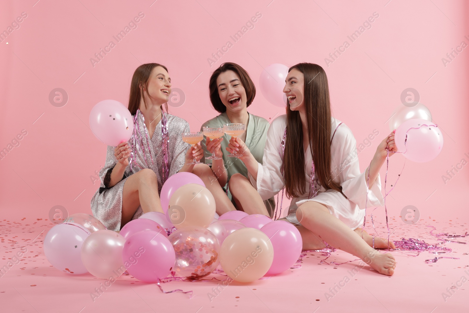 Photo of Pajama party. Happy friends with tinsel and balloons clinking glasses on pink background