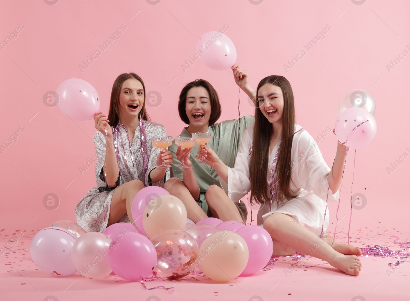 Photo of Pajama party. Happy friends with tinsel and balloons clinking glasses on pink background