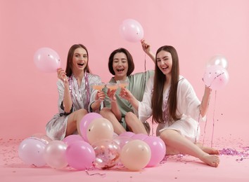 Photo of Pajama party. Happy friends with tinsel and balloons clinking glasses on pink background