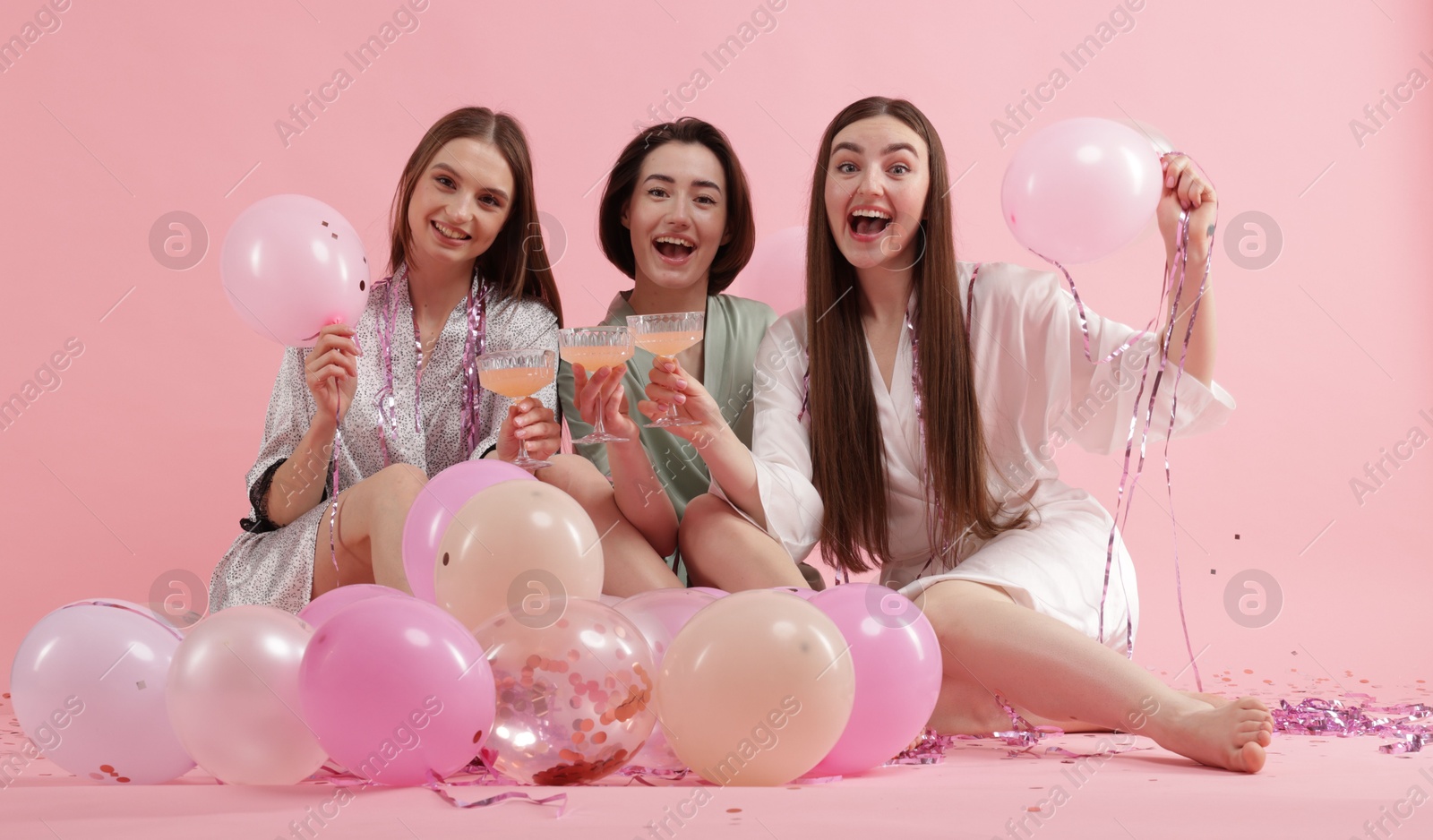 Photo of Pajama party. Happy friends with tinsel and balloons clinking glasses on pink background