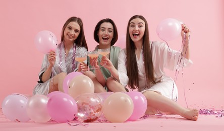 Photo of Pajama party. Happy friends with tinsel and balloons clinking glasses on pink background