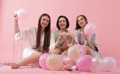 Photo of Pajama party. Happy friends with tinsel and balloons clinking glasses on pink background